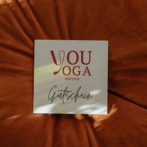 Quadratische Gutscheinkarte von "youyoga ROSTOCK" in hellem Design mit rotem Logo auf einem terracottafarbenen Samtkissen im Sonnenlicht.
