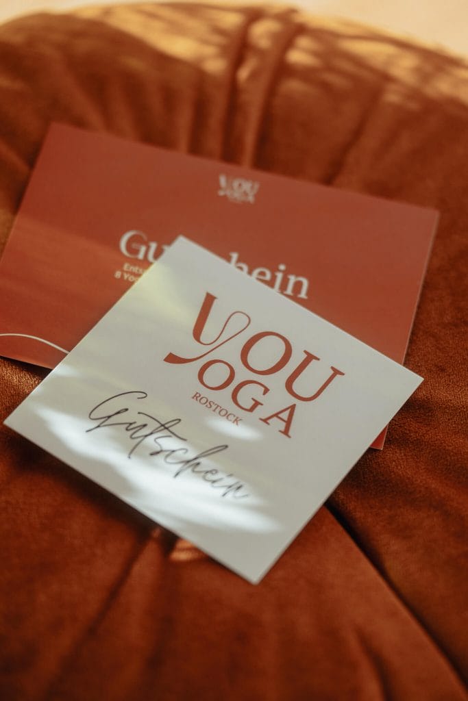 Zwei quadratische Gutscheinkarten von "youyoga ROSTOCK" in Weiß und Terrakotta auf einem samtenen, rötlichen Kissen.