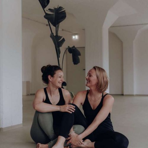 Die Yogalehrerinnen Julia und Johanna, eure Raumhalterinnen für das New Beginnings Retreat von youyoga, sitzen lächelnd in einem hellen Studio.