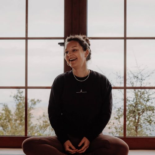 Kursleiterin Julia sitzt lächelnd auf einer Yogamatte vor einer Fensterfront während des Yoga und Fasten Retreats von youyoga Rostock.